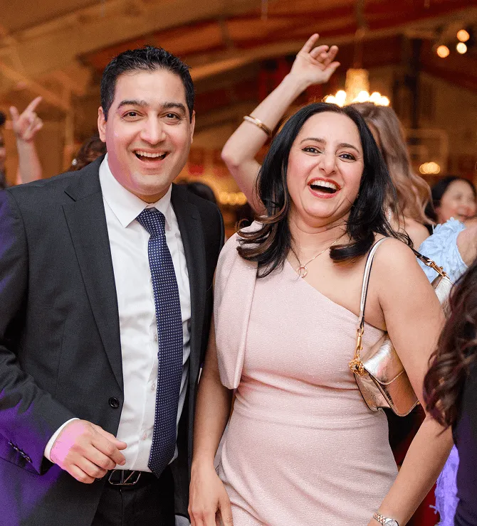 Couple smiling and enjoying a lively event, with festive atmosphere and guests in the background, representing joy and celebration.